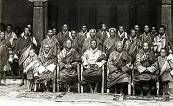 Ugyen Wangchuck, der Penlop von Trongsa, im Kreis seiner Ratgeber in Punakha, Bhutan, 1905. Vordere Reihe: Der Sohn von Thimphu Jongpen, Punakha Jongpen, Thimphu Jongpen, Trongsa Penlop, Zung Donyer [dronyer], Deb Zimpon und der ältere Sohn von Thimphu Jongpen. Alle Würdenträger sind in den gestreiften Roben gekleidet und sitzen vor einem Palastgebäude auf Stühlen.