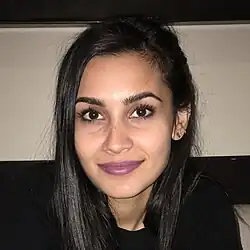 Sophia Ali mit langen, dunklen Haaren und einem freundlichen Lächeln. Sie trägt einen schwarzen Pullover. Auf dem linken Ohr ist ein kleines Ohrstecker-Piercing erkennbar. Der Hintergrund des Bildes scheint sich in einem Innenraum zu befinden. Eine beige Wand und ein dunkler, horizontaler Balken oder ein Regal bilden die Kulisse. Das Licht ist gedämpft, was darauf hindeutet, dass die Aufnahme bei schwachen Lichtverhältnissen gemacht wurde.