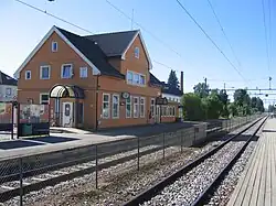 Foto eines Bahnhofsgebäudes mit Schienen im Vordergrund. Das Gebäude ist mit orange gestrichenem Holz verkleidet.