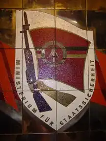 Wappenförmiges Emaille-Bild zeigt einen Arm, der ein Gewehr mit Bajonett hält, an dem eine rote Fahne mit den Symbolden der DDR befestigt ist