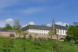 Oberhalb einer Streuobstwiese und einer roten Sandsteinmauer erhebt sich das weiße Stift mit der Kapelle.