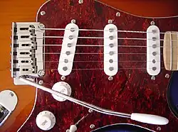 Detailbild der Vorderseite des Korpus einer rechtshändigen Squier Stratocaster in Sunburst-Lackierung mit einem Pickguard in rotbraunem Schildpattmuster. Rechts im Bild der Ansatz des ahornfarbenen Griffbretts, links Steg und Vibratohebel in Chromoptik, dazwischen drei Einzelspulentonabnehmer in weißer Umrahmung, darunter im Bild jeweils leicht nach links versetzt der weiße teilweise verdeckte Drehregler mit der erkennbaren Aufschrift „VOL“ (Rest verdeckt) sowie ein weißer Drehregler mit Aufschrift „TONE“, darunter nach rechts versetzt ein Schiebeschalter mit weißer Kappe. In der linken unteren Bildecke ist knapp eine chromfarbene Klinkenbuchse erkennbar.