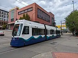Straßenkreuzung mit zweigleisiger Straßenbahnstrecke und dreiteiligem Straßenbahn-Triebwagen in Bildmitte. Den Hintergrund dominiert ein in dunklen, teils schmutzigen Rottönen gehaltenes modernes viergeschossiges Geschäftsgebäude. Der Triebwagen ist weiß und hat im unteren Viertel auf ganzer Länge ein stilisiertes Meereswellen-Muster in Blau- und Grüntönen. Die festen Anschriften weisen in Verbindung mit Tacoma-Link-Logos auf die Marke TACOMA LINK und den Betreiber SOUND TRANSIT hin. Auf dem Zielschild steht ST JOSEPH.