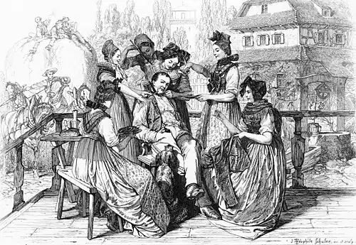 Auf einer Terrasse sitzt ein junger Mann lässig auf einem Stuhl; sechs in Trachten gekleidete Frauen umgarnen ihn.