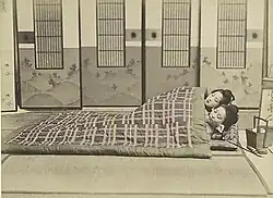 Zwei Personen liegen auf einem Futon und sind mit einer Decke bedeckt. Das Bild ist in sepia.