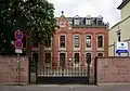 Thebäerstraße 51: ehemaliges Schulhaus des Vororts St. Paulin; siebenachsiger sogenannter Rohbau: Stichbogenfenster und Brüstungsfelder hell abgesetzt vom kleinteiligen Sandsteinquadermauerwerk, 1872, Architekt A. Massing, 1884 verlängert