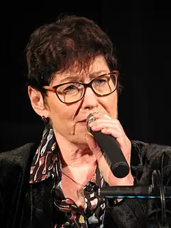 Uschi Brüning ist ein Mikrofon halten au einer Farbfotografie im Hochformat abgebildet. Sie hat kurzes braunes Haar und trägt eine Brille.