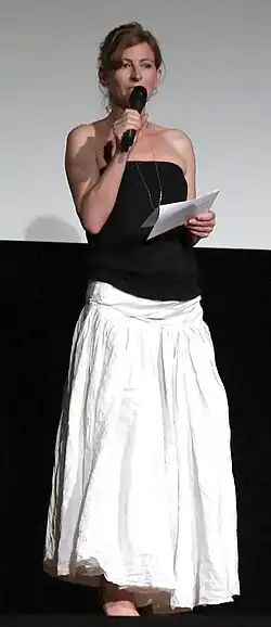 Tatjana Alexander steht sprechend auf der Bühne, der Preisverleihung der Vienna Independent Shorts 2014. Sie hält in einer Hand ein Mikrofon, während sie in der anderen Hand ein Blatt Papier hält. Tatjana Alexander trägt ein schwarzes schulterfreies Oberteil und einen langen, weißen Rock. Im Hintergrund ist eine schlichte Wand zu sehen, die im unteren Bereich schwarz ist.