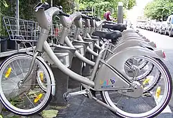 Seitliche Farbfotografie von einer Reihe silberner Fahrräder derselben Marke mit Körben vor den Lenkrädern, die an Ladestationen stehen. Am Hinterrad steht in bunter, kurviger Schrift „Velib’“ und darunter „Mairie de Paris“. Im Hintergrund betätigt ein Mann einen Automaten.