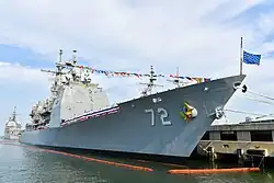 Über die Toppen geflaggter Kreuzer an Pier aus Richtung Wasser aufgenommen, Bug voraus. Kennnummer 72 zentral im Bild. Flagge Navy Jack, tiefblau mit den goldenen Sternen der Bundesstaaten, ist auf dem Vordersteven halb gehisst. Schiff ist von Ölbarrieren umgeben und liegt hoch im Wasser. Im Hintergrund der Achtersteven eines Schwesterschiffes und Masten anderer Schiffe.