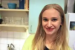 Vera Vitali Junge Frau mit langen, blonden Haaren und lächelnden Gesicht, die in einer modernen Küche steh lächelndem Gesicht, trägt ein helles Oberteil, steht in einer Küche. Die Haare fallen locker über die Schultern, und sie trägt ein hellgrünes Oberteil über einem schwarzen Shirt.Die Küche ist mit hellen Schränken ausgestattet, die glatte Fronten haben. Eine weiße, geflieste Rückwand sorgt für eine saubere und helle Atmosphäre. Auf den Regalen stehen verschiedene Gegenstände wie Gläser und Schüsseln.Links unten im Bild ist ein Waschbecken mit einer Armatur sichtbar. Im Hintergrund sieht man einen Kühlschrank, an dessen Tür ein kleines Bild oder Magnet angebracht ist.Die gesamte Szene vermittelt den Eindruck eines gemütlichen und funktionalen Küchenbereichs in einer privaten Wohnung.