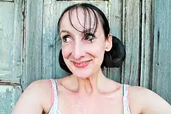 Viola vor einer verwitterten Holzwand. Sie hat dunkles Haar, das zu zwei Knoten gebunden ist und einen dünnen Pony. Sie lächelt und blickt zur Seite. Ihr Oberteil hat dünne Träger und ein helles Muster. Die Holzwand im Hintergrund ist blaugrün und zeigt Abnutzungsspuren.