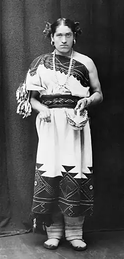 Schwarz-Weiß-Foto von We’wha. We’wha trägt ein traditionelles, helles Gewand, das bis zu den Unterschenkeln reicht und den gesamten linken Arm freilässt. Auf dem Gewand befinden sich mehrere Muster, unter anderem schwarze Dreiecke und weiße Linien auf schwarzem Hintergrund. Am rechten Ärmel und dem Ende des Gewands hängen weiße und schwarze Bommel. We’wha trägt daneben runde Ohrringe, eine bis zur Brust reichenden Steinchen-Halskette mit drei Ringen am Ende, einen schwarzen Gürtel mit weißen Rauten und schwarzen Dreiecken, unter dem Gewand eine bis zu den Füßen reichende graue Hose und pantoffel-ähnliche Schuhe. In der linken Hand hält We’wha ein weißes Körbchen mit schwarzen Linien. An den Seiten von We’whas kurzen, bis zu den Ohren geschnittenen schwarzen Haaren befinden sich lockenartige Kringel.
