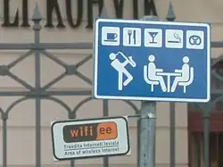 Farbfotografie von Straßenschildern mit einem Eisenzaun und einer Häuserfassade im Hintergrund. Ein blau-weißes Schild zeigt zwei Figuren an einem Tisch und einen Musiker, der links im Stehen spielt. Über ihnen sind kleine Symbolfelder für Café, Restaurant, Bar, Kiosk, Bäckerei. Das kleinere Schild zeigt das Symbol für Wifi mit dem Text „Area of wireless internet“.