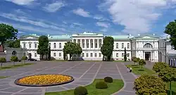 Frontale Farbfotografie eines weißen, zweigeschossigen Gebäudes mit grünem Dach. Im mittleren Obergeschoss bilden Säulen eine Loggia. Vor dem Gebäude ist ein großer Platz mit einem runden Beet mit gelben Blumen. Am Rand sind kleine Rasenflächen und einige junge Bäume.