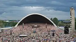 Farbfotografie in der Obersicht von einer riesigen Menschenmenge, die eine Open-Air-Bühne bis zur Konzertmuschel ausfüllt. Mehrere Lautsprecheranlagen sind verteilt und rechts steht ein grauer, langer Turm. An beiden seitlichen Mauern stehen aufgereiht blau-schwarz-weiße Nationalflaggen. Im linken Hintergrund ist der Hafen mit Schiff zu sehen.