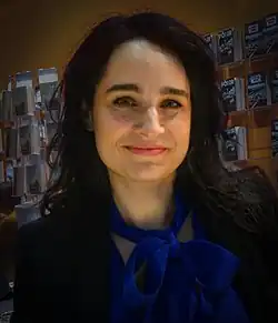 Yael Adler (2016) lächelt freundlich mit langen, dunklen Haaren und in einem professionellen Outfit: ein dunkler Blazer und eine blaue Bluse mit einer großen Schleife am Hals. Im Hintergrund sind mehrere Zeitschriften oder Broschüren auf Regalen ausgestellt, was auf einen Innenbereich hindeutet. Es könnte sich um eine Buchhandlung, Bibliothek oder eine Veranstaltung zur Präsentation von Publikationen handeln.