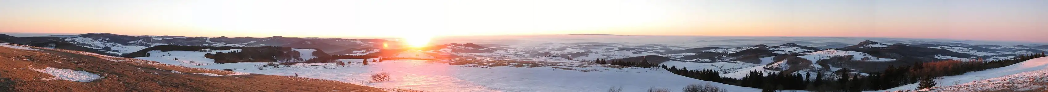 Sonnenuntergang unterhalb des Radoms. Hier sind Tische und Bänke vorhanden, von denen aus man den Westblick mit Sonnenuntergang genießen kann. Zum Panorama: Aufgrund einer Inversionswetterlage herrscht sehr gute Fernsicht. Der Berg mit Funkturm ganz links im Bild ist der Kreuzberg. Rechts neben der Sonne liegt am Horizont der Vogelsberg (Entfernung etwa 45 km).  Zwischen Sonne und Vogelsberg erkennt man in der Bildervergrößerung übrigens sogar den Grossen Feldberg im Taunus (Luftlinie etwa 110 km). Rechts im Vordergrund liegt die Kuppenrhön mit der markanten Erhebung der Milseburg. Noch weiter Rechts ist am Horizont ein länglicher Höhenzug zu erkennen. Dies ist der Hohe Meißner (Entfernung etwa 80 km).