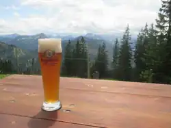 Im Vordergrund ein Weißbier im Glas auf einer Biertischgarnitur, im Hintergrund Bergpanorama